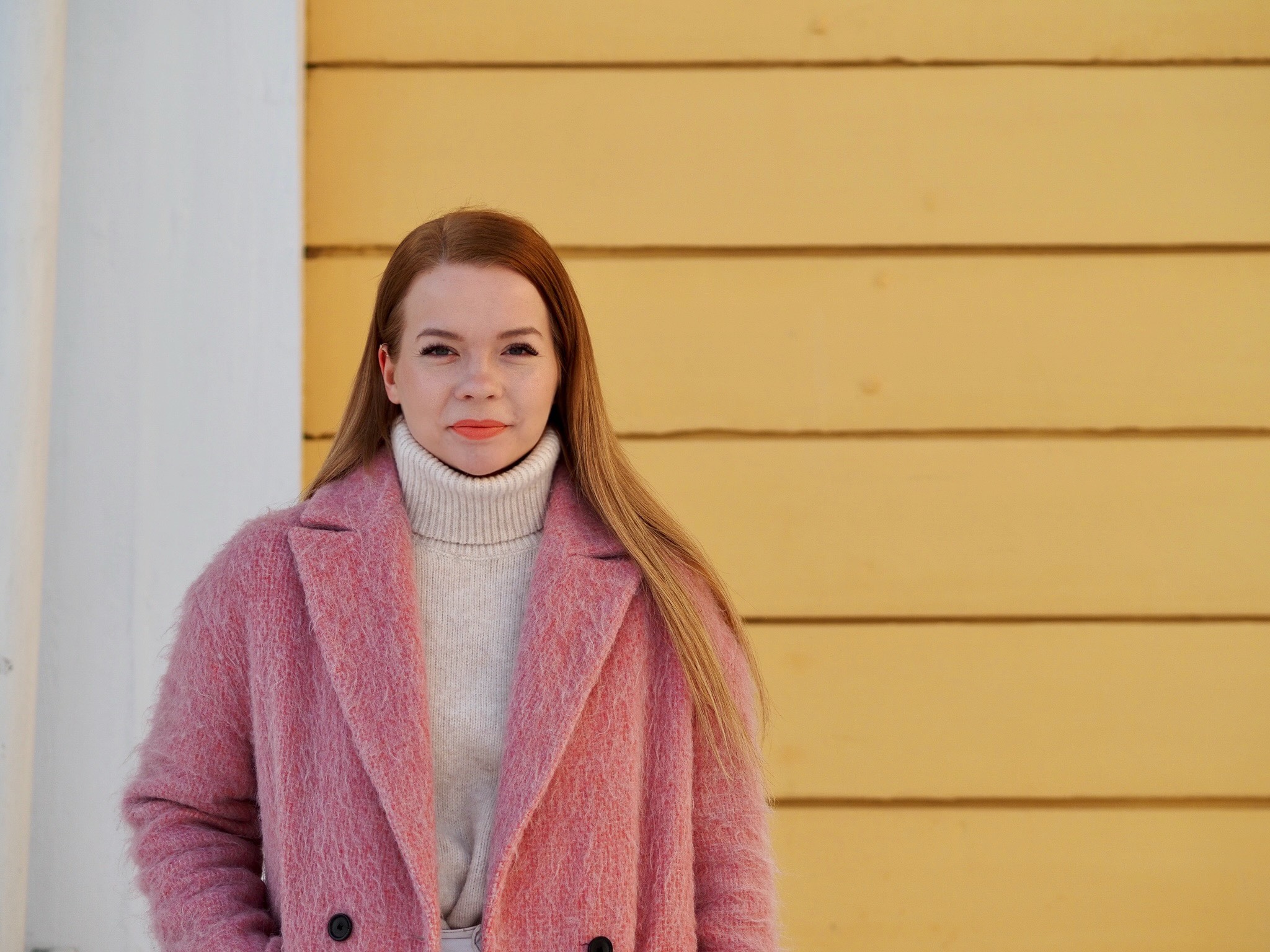 Kuvassa tuottaja Roosa Yrjölä keltaisen seinän edessä pinkissä takissa
